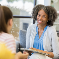 photo of a black, female counselor smiling empathetically at a student while holding a clipboard and pen