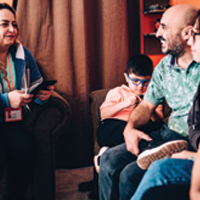 photo of casually dressed in-home care provider wearing a name tag and holding a notebook and pen speaks to a couple and their two children
