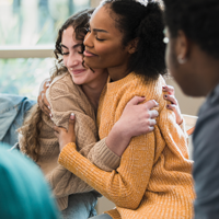 photo of white female high school student and black female high school student sitting in a circle of chairs hugging while a black male high school student looks on